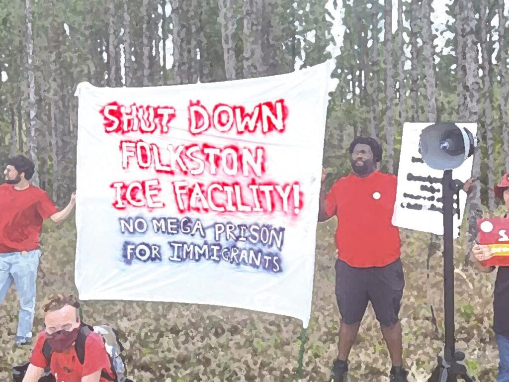 A group of protesters in red shirts stand in front of a wooded area holding a large white banner that reads “Shut Down Folkston ICE Facility! No Mega Prison for Immigrants.”