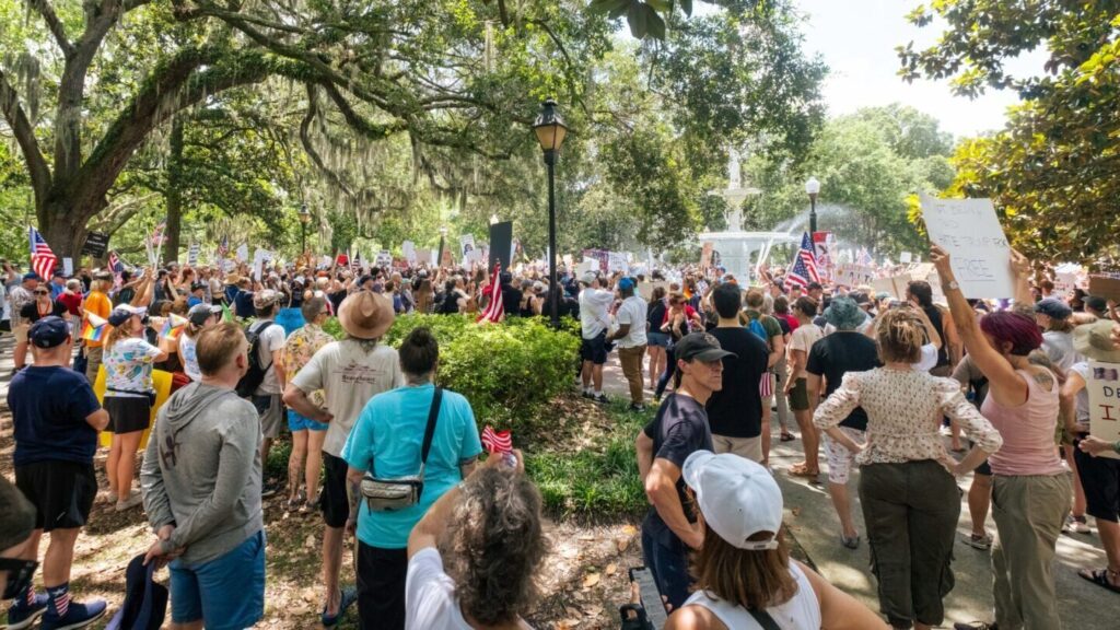 A wide-angle, slightly elevated shot of a large crowd of protesters gathered around the iconic white fountain in Forsyth Park, Savannah, Georgia. The scene is on a sunny day, with large trees draped in Spanish moss framing the rally.