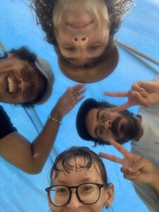 Four people lean over the camera and smile under a blue tarp in the rain, with two of them flashing peace signs.