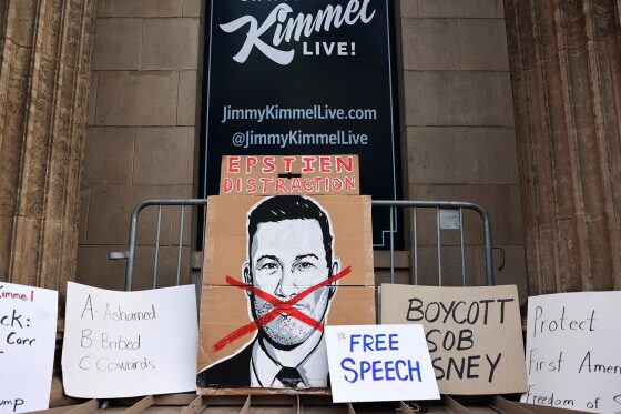 A protest outside a "Kimmel Live!" studio, featuring multiple handmade signs. A large cardboard sign in the center displays a black and white portrait of a man with a red 'X' over his face. Above this, a red sign reads "EPSTEIN DISTRACTION". Other signs include "A: Ashamed B: Bribed C: Cowards," "BOYCOTT SOB SNEY," and "FREE SPEECH.