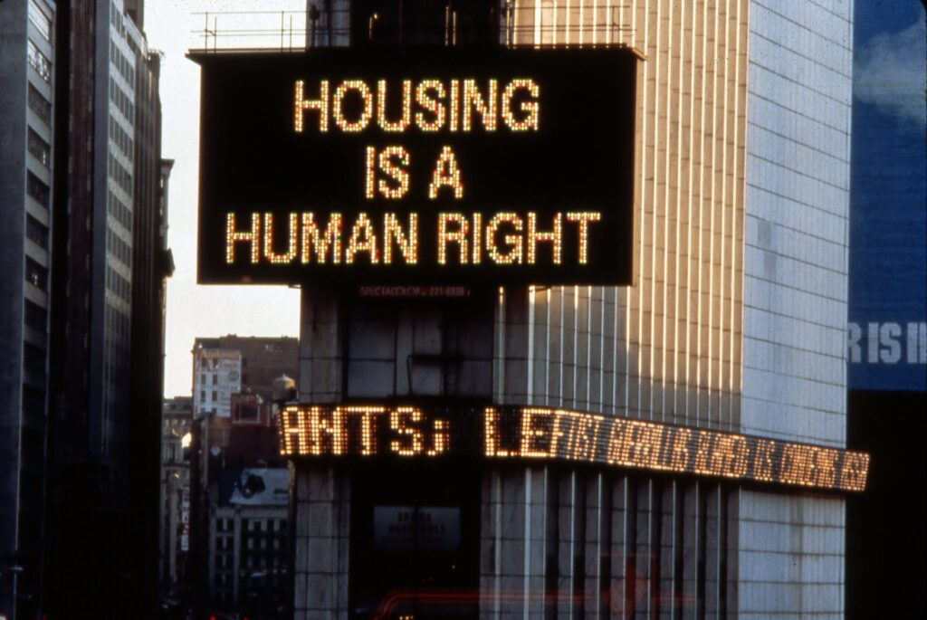 Large electronic Spectacolor billboard in New York’s Times Square reading “HOUSING IS A HUMAN RIGHT,” mounted on a tall building above the city streets.