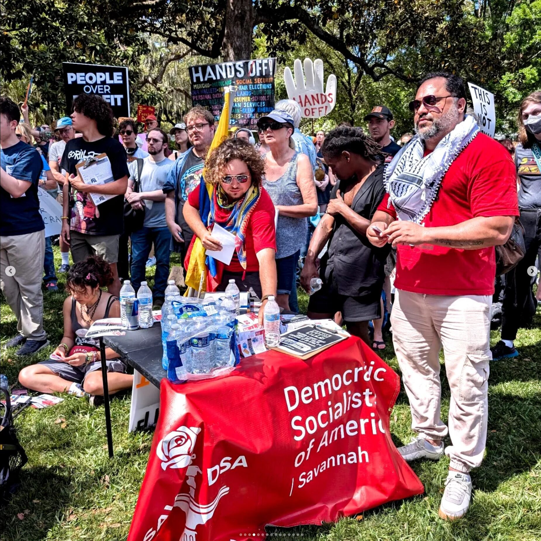 People with a red “Democratic Socialists of America | Savannah” banner table at a crowded outdoor rally, surrounded by protesters holding signs like “People Over Profit” and “Hands Off.”