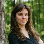 A Caucasian woman with long brown hair standing outdoors beside a tree, wearing a black V-neck top, with a soft green forest background.