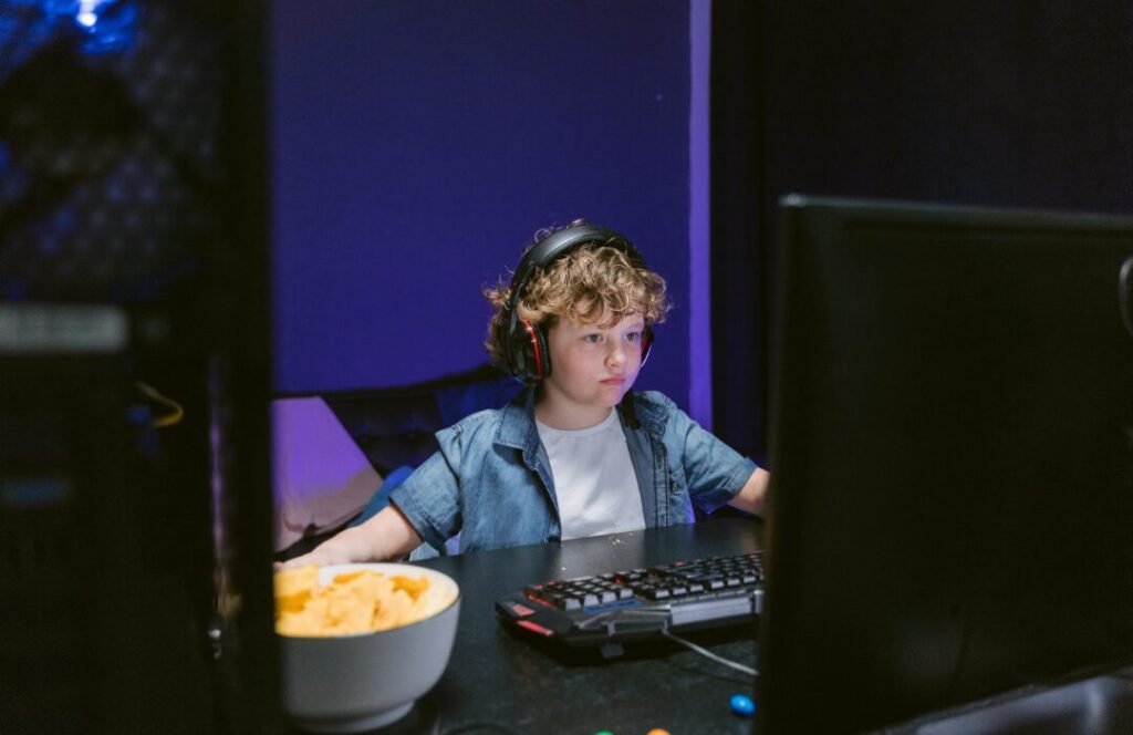 A young boy with curly brown hair and headphones sits intently in front of a computer screen, a bowl of chips beside him, in a room with purple lighting.