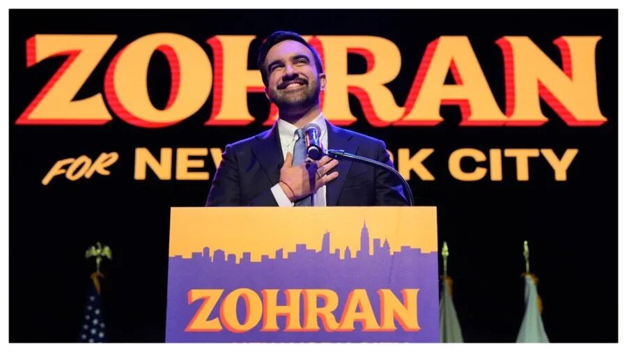 A smiling Zohran Mamdani stands at a podium labeled “ZOHRAN,” in front of a large campaign-style backdrop reading “ZOHRAN for New York City.”