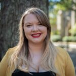 Kendra Clark, Caucasian, smiling, standing outdoors in front of a tree, wearing a yellow cardigan over a black top, with soft, blurred greenery and buildings in the background.
