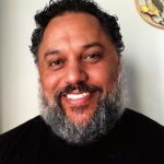 George Robles, Hispanic, smiling, standing indoors with a plain white wall behind. He is wearing a black shirt, with "Free Palestine" visible.