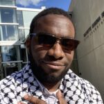 Keyshawn Housey, African American, smiling, Outdoors in front of a building. He is wearing a light colored button-up shirt with a black and white keffiyeh draped over his shoulders